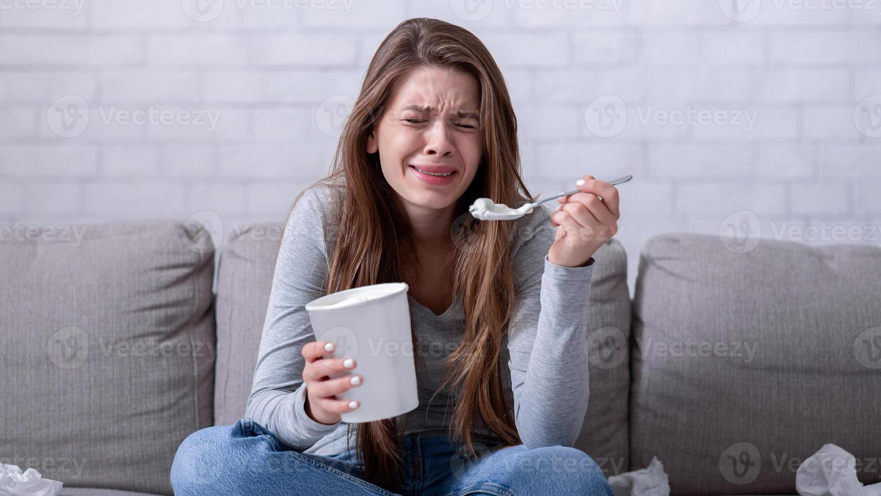 Emotional eating concept. Stressed millennial lady crying on sofa with bucket of ice cream, suffering from relationship problem or watching sad movie, feeling lonely and depressed photo
