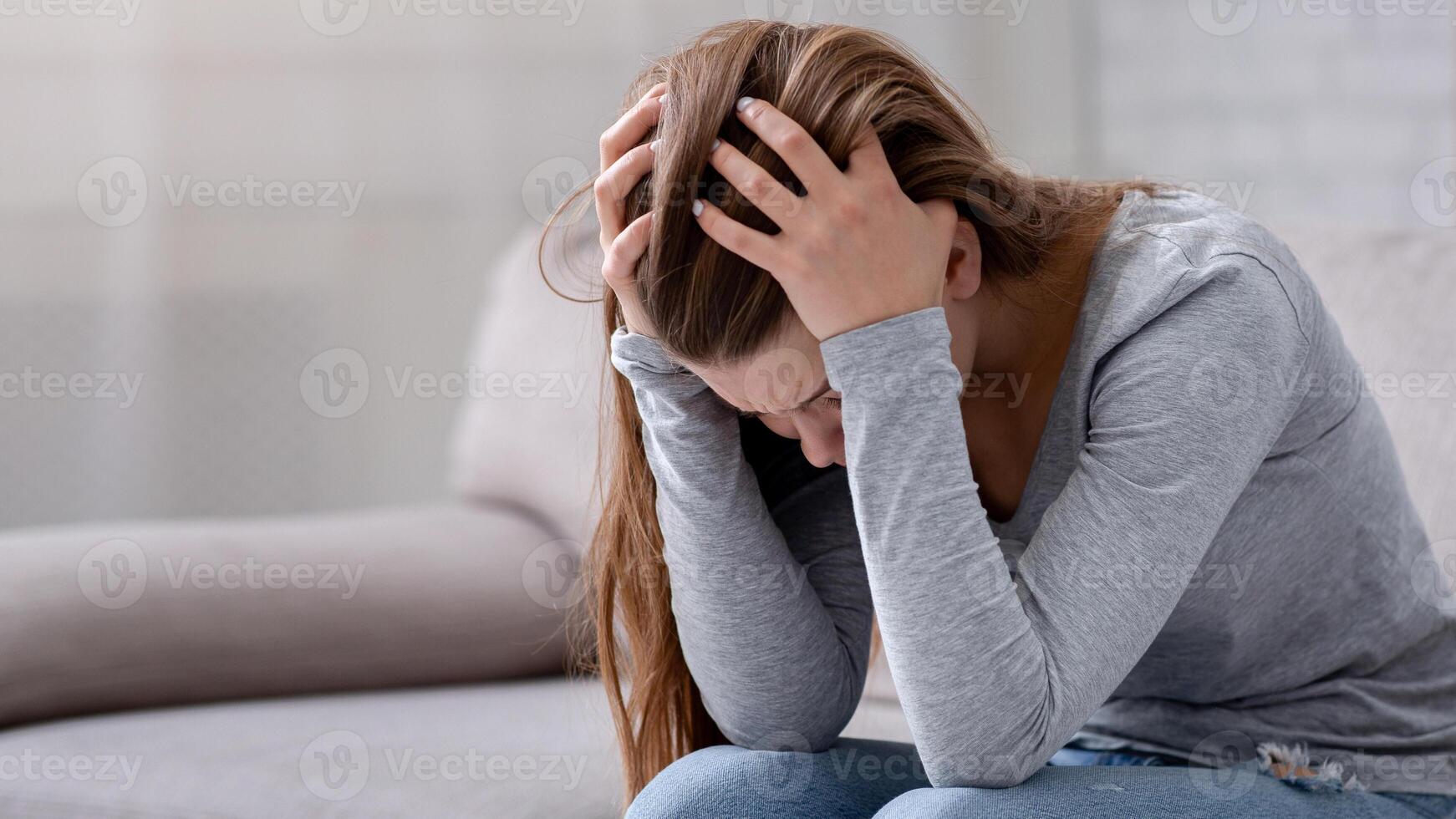 Unhappy young woman with head in her hands sitting on couch, crying and feeling depressed. Desperate millennial lady suffering from mental problems, broken heart or mood disorder photo