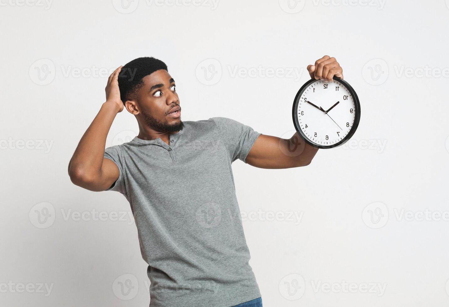 Punctuality and time management concept. Shocked young african-american man looking at wall clock and holding his head, white background photo