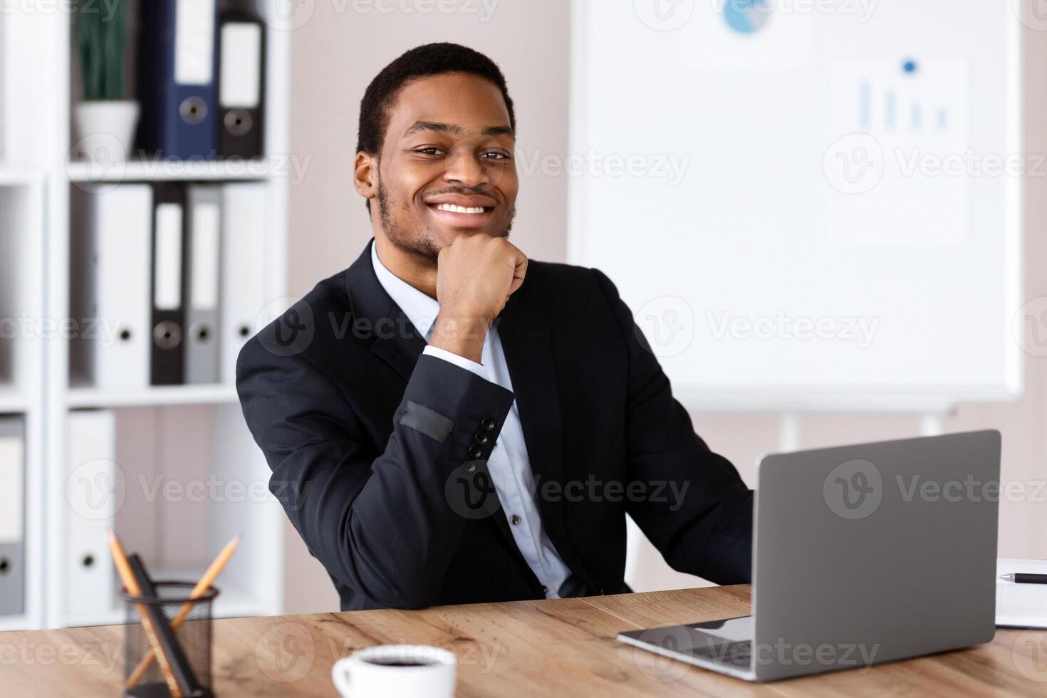 Successful african american businessman sitting in his office, using modern laptop, drinking coffee, smiling at camera, copy space. Cheerful black entrepreneur starting his day with reading emails photo