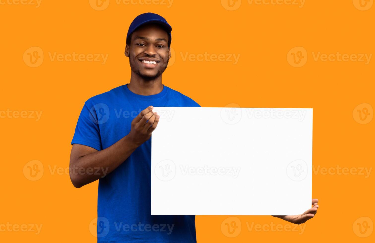 Shipment Service Promotion. Portrait of smiling bearded black delivery man in blue uniform holding and showing square empty blank board with template isolated on orange studio background photo