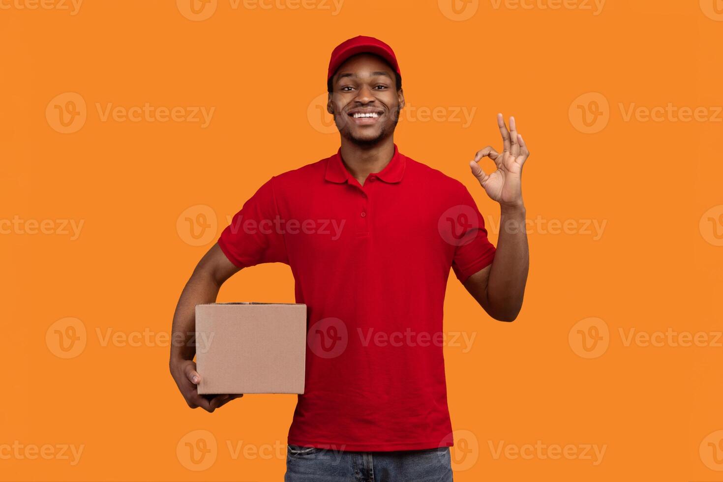 Best Delivery Service Concept. Smiling black male courier wearing red cap and uniform holding cardboard box and showing okay sign gesture standing isolated over yellow orange studio background photo