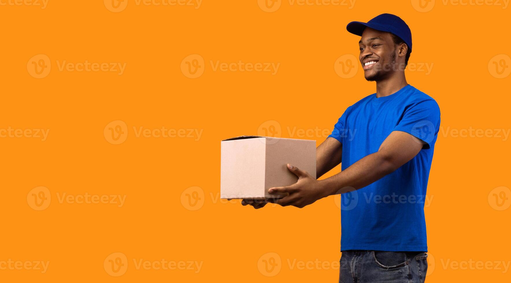 Delivery Service Concept. Smiling black male courier wearing blue cap and uniform holding cardboard box, showing and giving it to the side, copy space, banner. Guy standing on yellow studio background photo