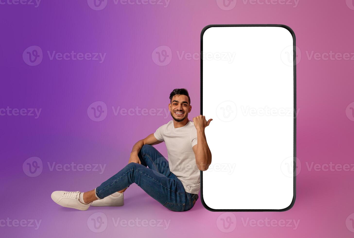 Full length of cheerful young Arab man sitting near big cellphone with mockup for mobile app on screen in neon light. Smartphone display template for new website or online ad photo