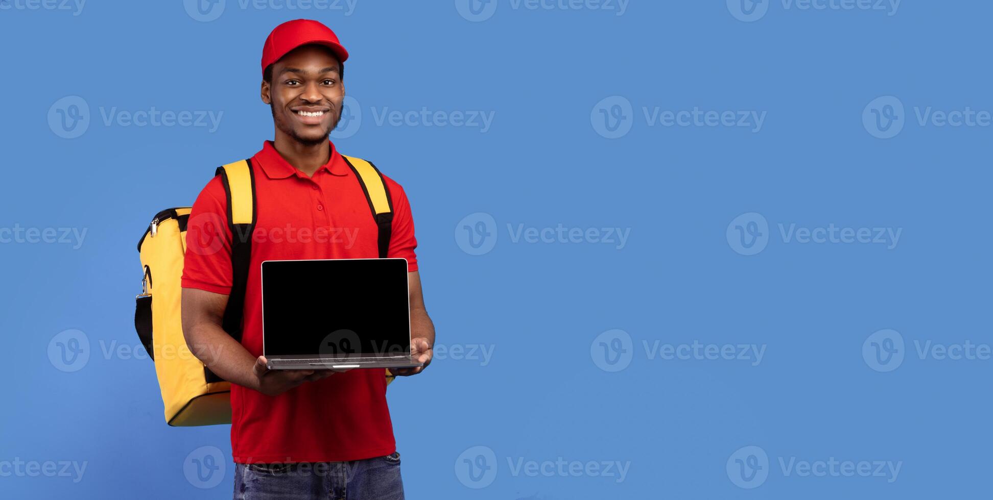 African american courier man holding laptop with blank screen for mock up, wearing red uniform and thermo backpack bag, standing isolated on blue studio background, panorama, banner, copy space photo