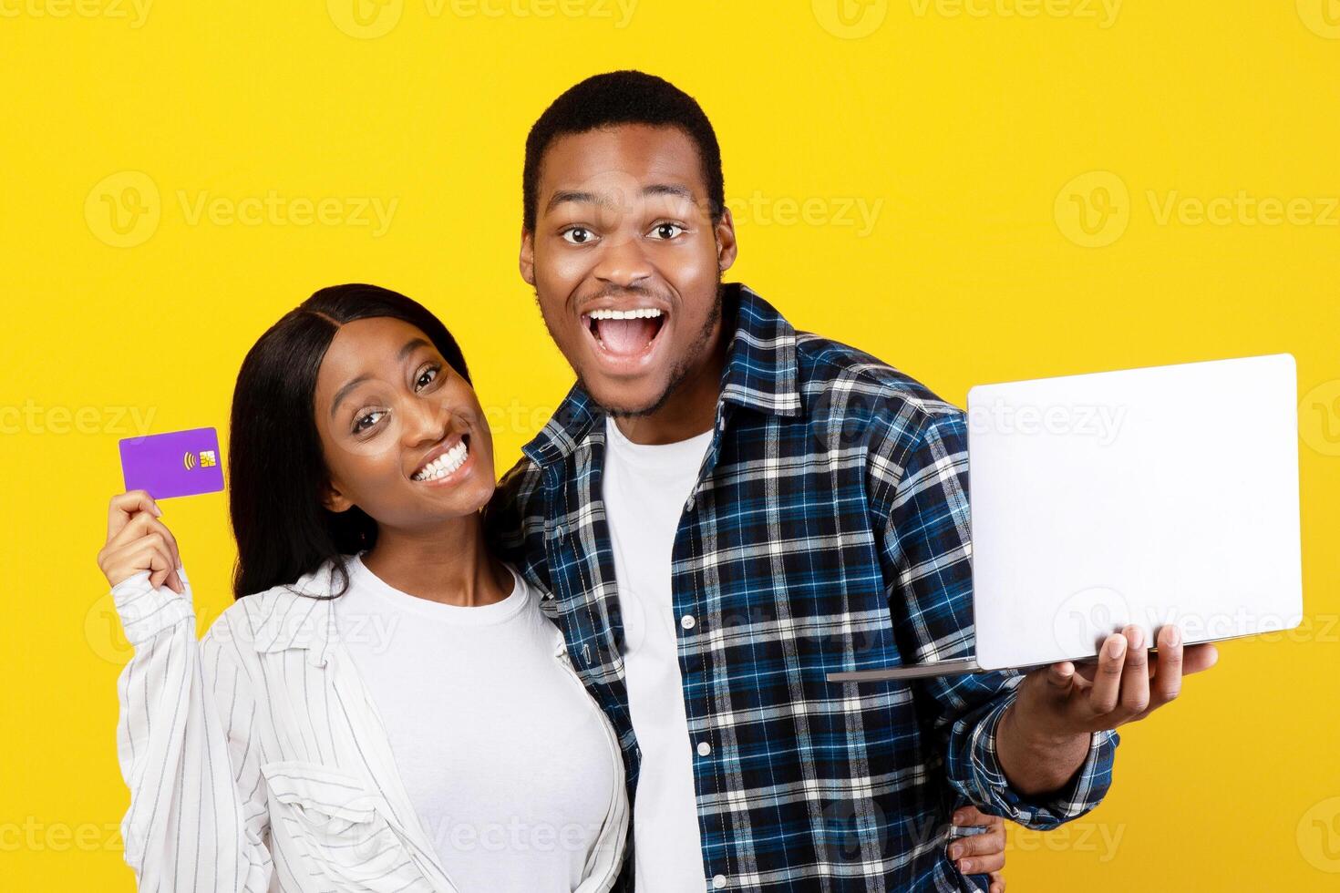 Online shopping, black friday and payment with money. Happy laughing young african american guy with open mouth holds laptop, female shows credit card, isolated on yellow background, studio shot photo