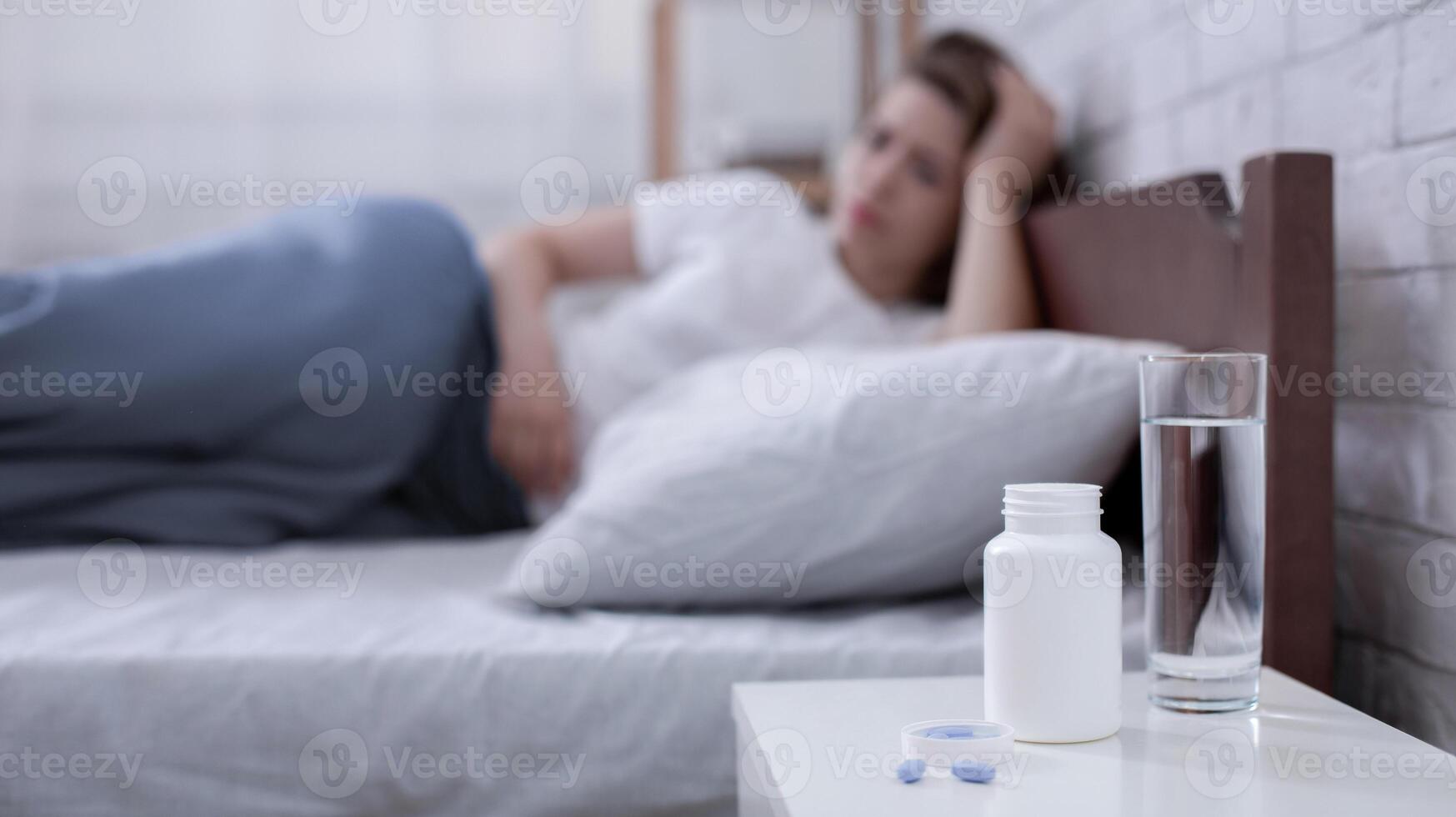 Jar of pills and glass of water on bedside table and young depressed woman lying on bed in background, copy space. Selective focus. Suicide and sleep deprivation problem concept photo