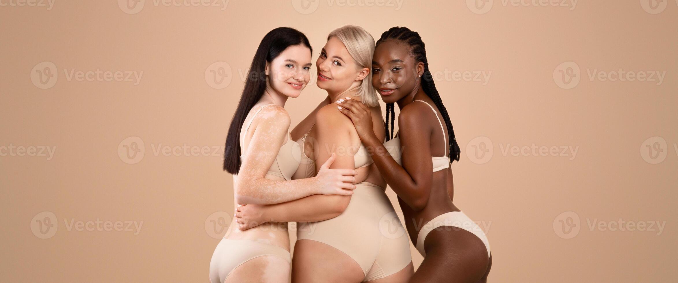 All bodies are good bodies. Three happy body positive ladies posing together in lingerie over beige studio background, diverse females standing half-turned at camera and embracing, copy space photo