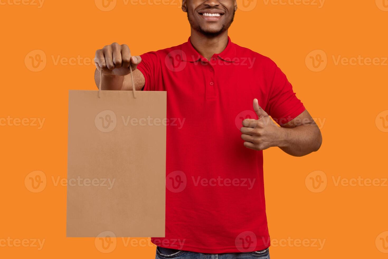 Closeup of unrecognizable black man in red t-shirt holding craft paper bag, showing thumbs up gesture sign, isolated on orange background. Design, mockup, blank space on package for advert photo