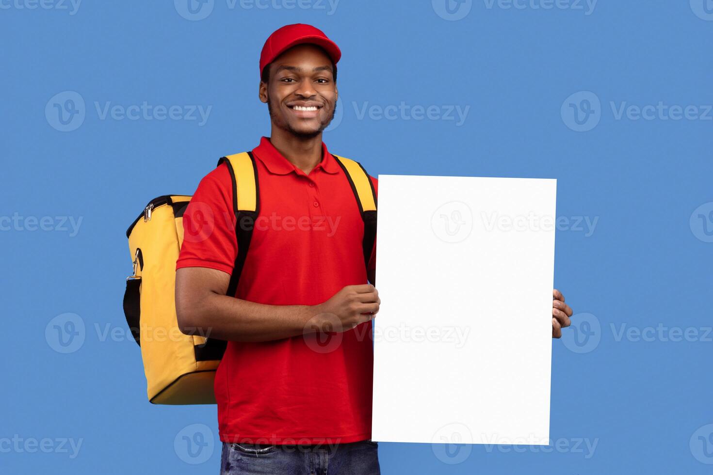 Shipment Service Promotion. Portrait of cheerful bearded black delivery man wearing thermo bag holding and showing vertical empty blank board sign standing isolated on blue studio background photo