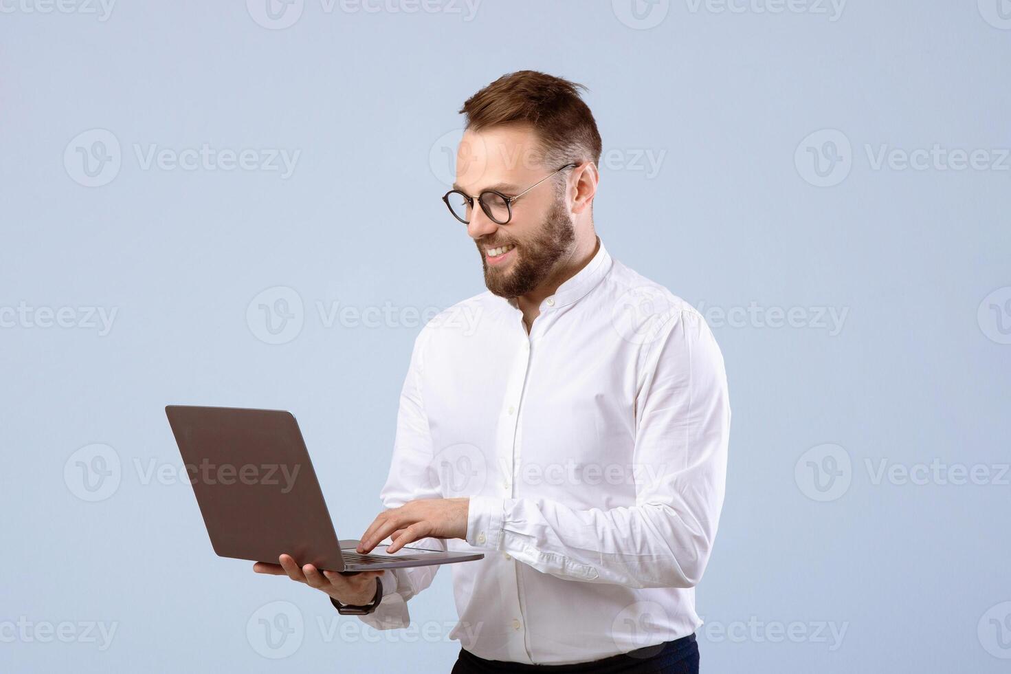 Positive young man in office wear using laptop computer over grey studio background. Joyful millennial guy working or studying remotely, having online business meeting on pc photo