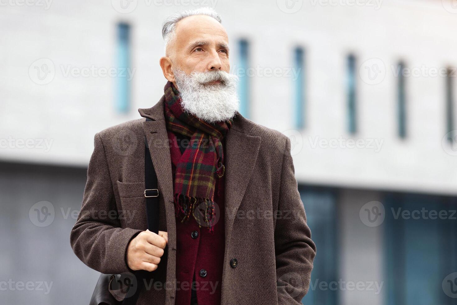 Thoughtful senior grey-haired man in stylish burgundy jacket walking by street alone, going home from job, looking at copy space. Working during retirement concept, active lifestyle of elderly photo