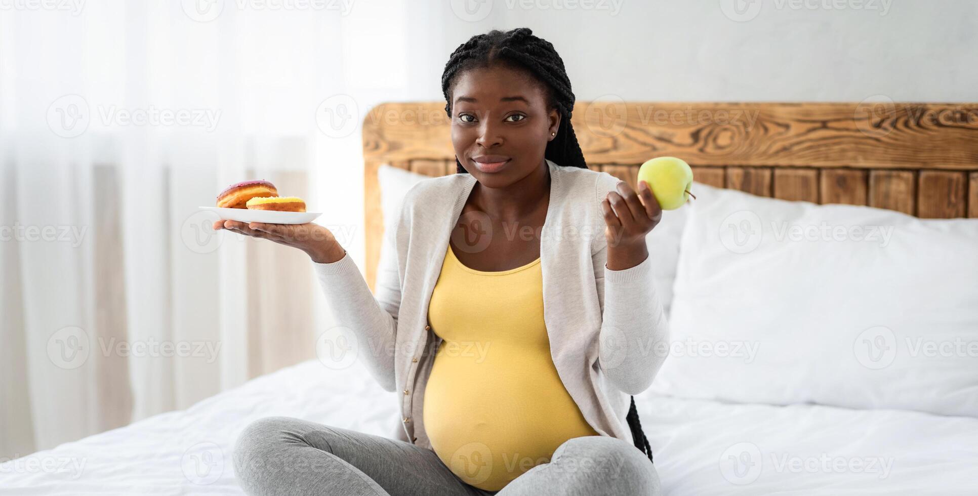 Pregnant black woman choosing between healthy and unhealthy food at home, doubtful expectant lady sitting on bed holding green apple and plate with glazed donuts, looking at camera, copy space photo
