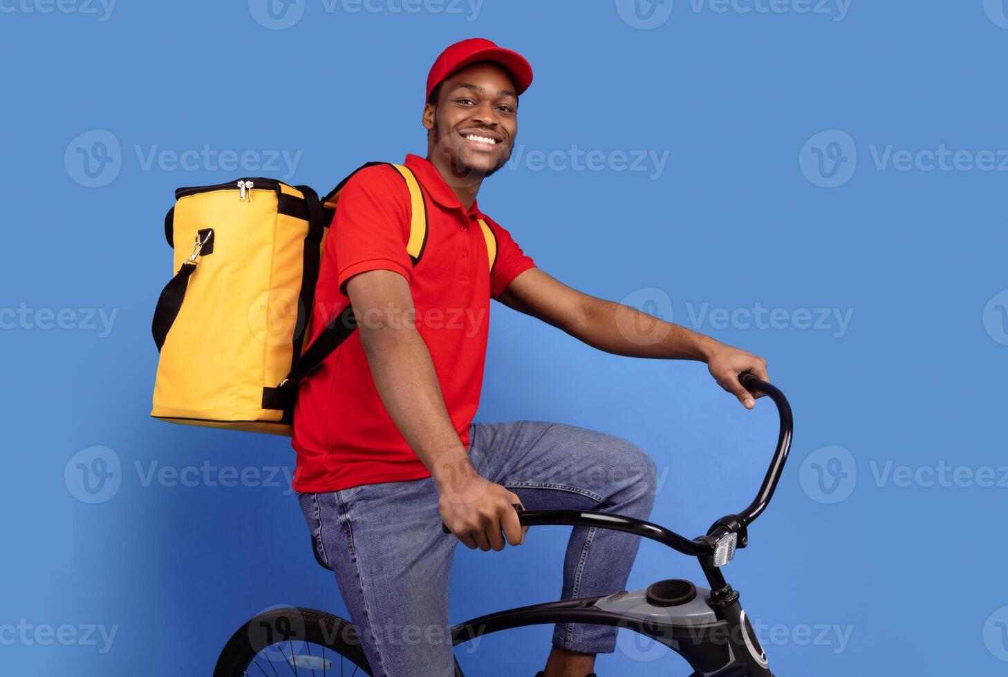 Fast Delivery Service. Smiling african american courier guy in red cap and uniform riding bicycle and wearing yellow thermo backpack bag isolated on blue studio background. Shipment and post photo