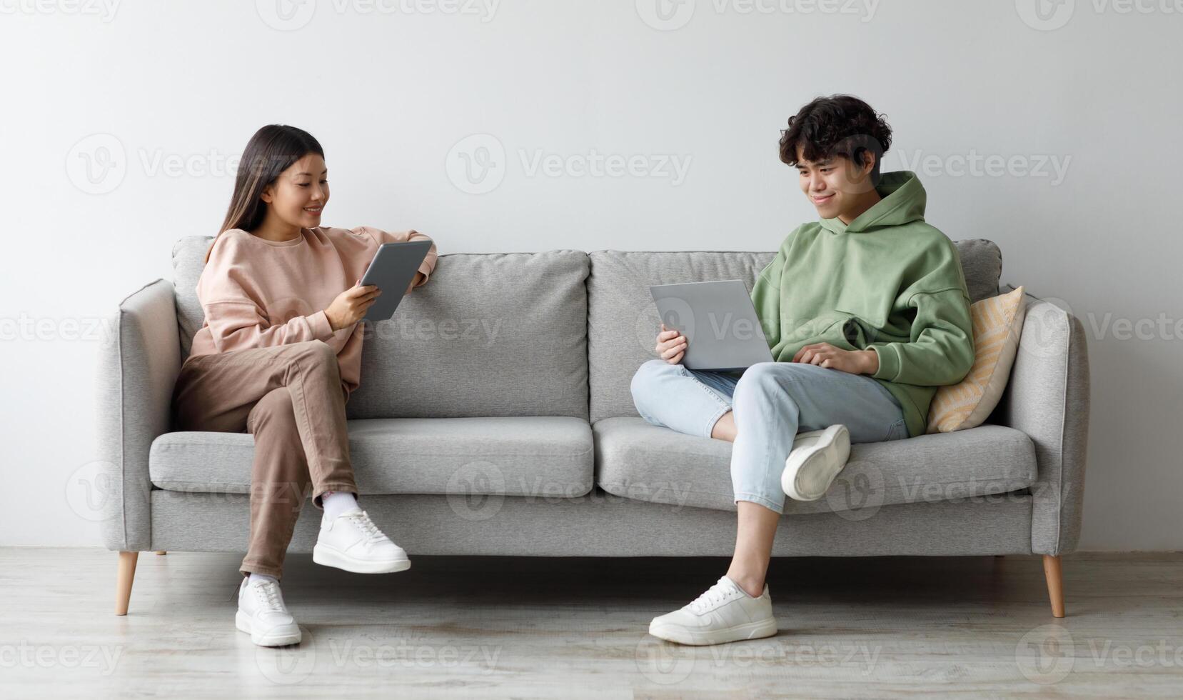 Handsome young Asian guy and his girlfriend using modern gadgets while sitting on couch at home, copy space. Full length of millennial couple communicating online via laptop and tablet pc photo