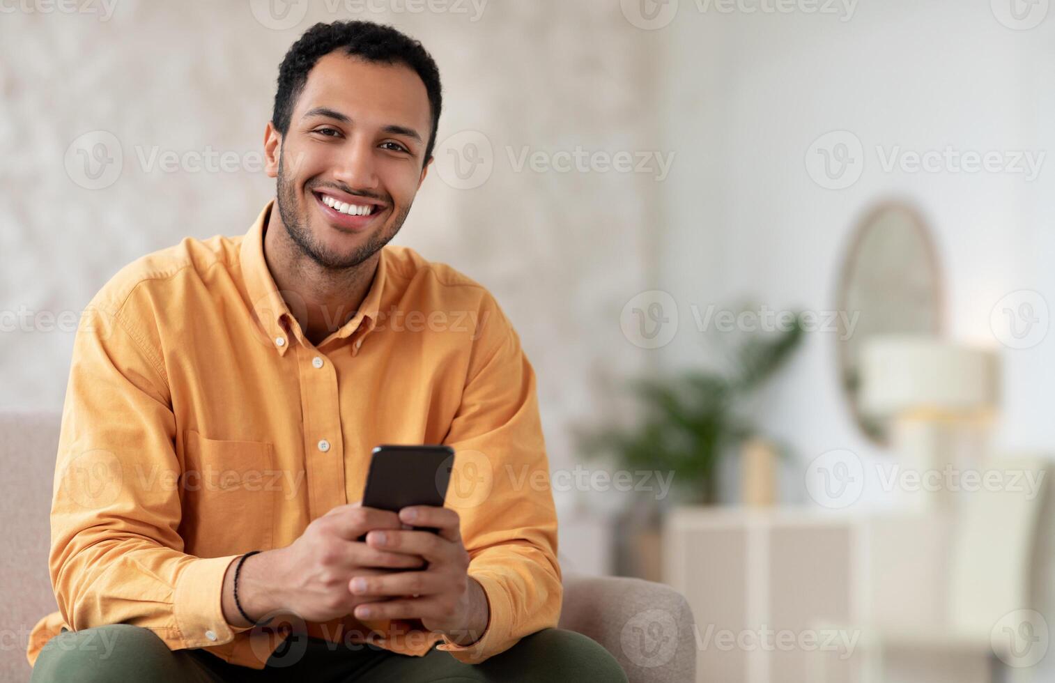 Happy Smiling Man Using Phone Browsing Internet Sitting On Couch At Home. Cheerful Cellphone User Testing New Application For Smartphone, Posing Looking At Camera, Free Copy Space, Mockup photo