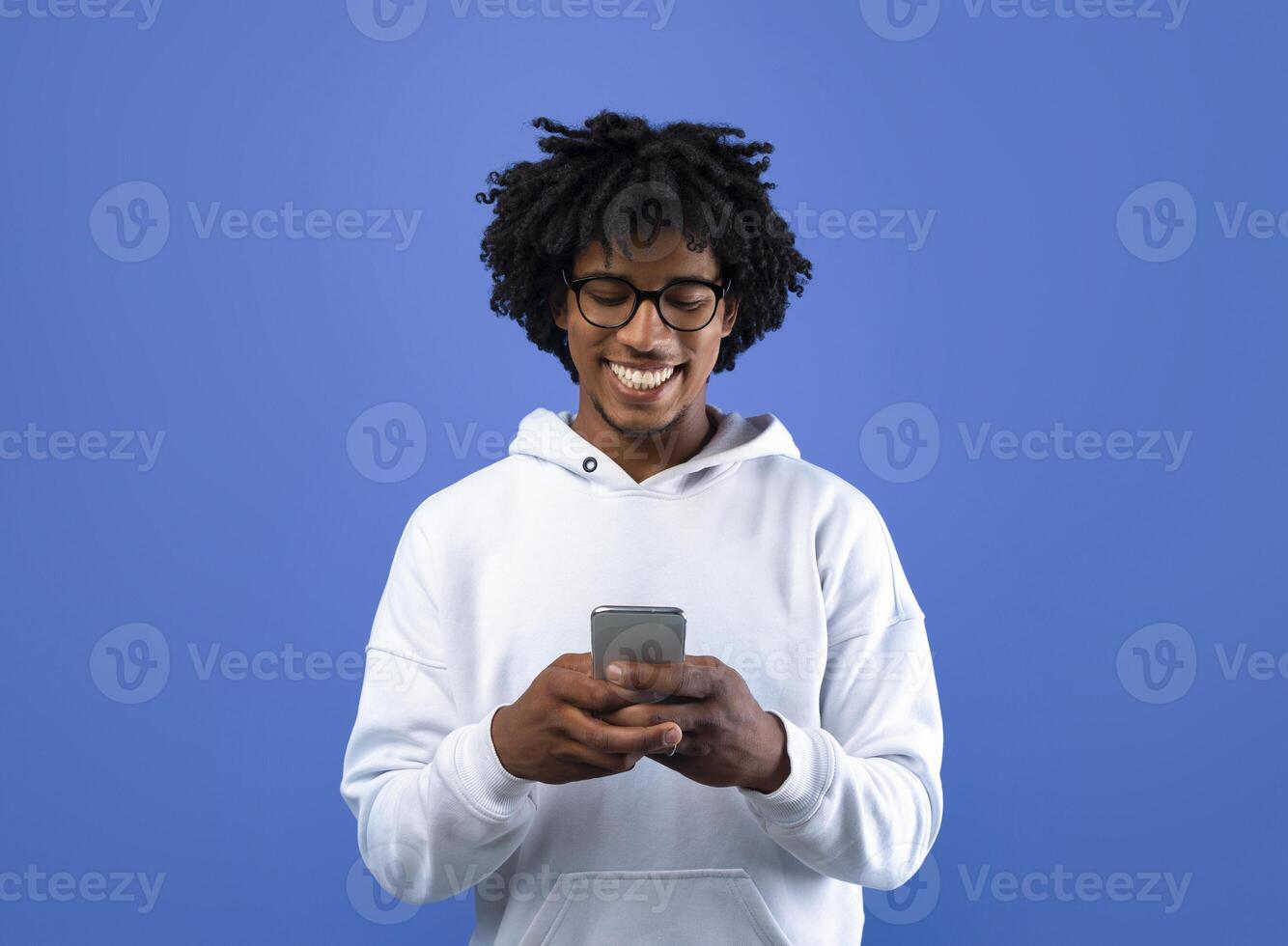 Happy black teenager looking at mobile phone screen, using new cool application on blue studio background. Joyful African American adolescent using smartphone, browsing social network photo