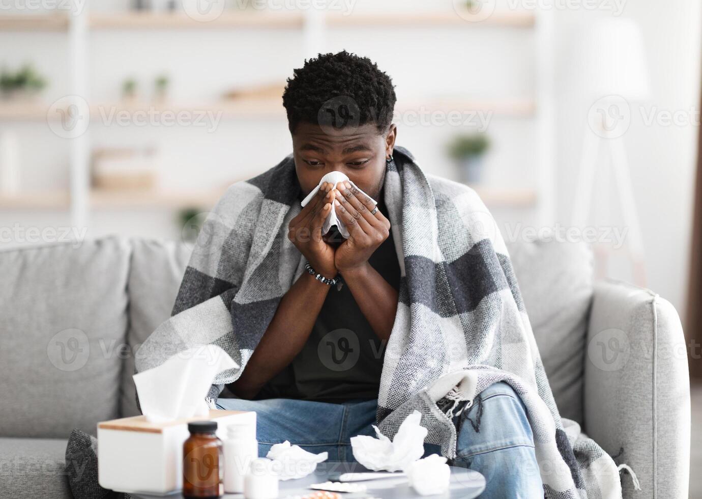 Sick black guy covered in blanket sitting on couch and sneezing, using napkins and medicine, got sick while COVID-19 pandemic, home interior, copy space. Cold, flu, coronavirus concept photo