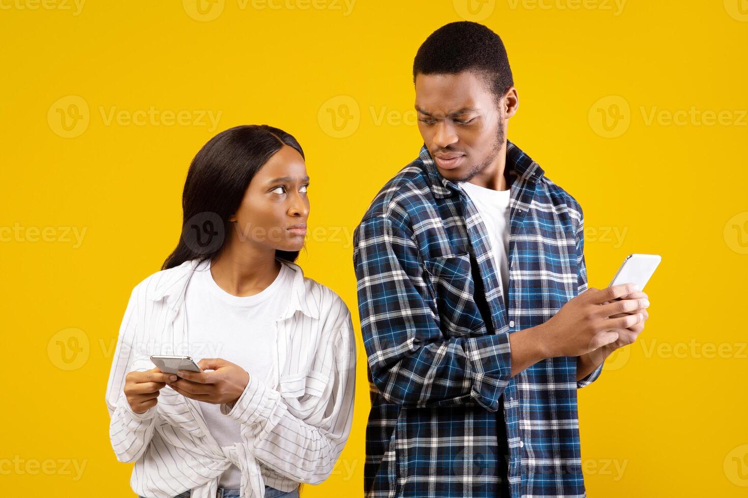 Relationship, problems and modern devices. Upset african american husband and wife look at each other with anger and disbelief and hold phones, isolated on yellow background, studio shot photo