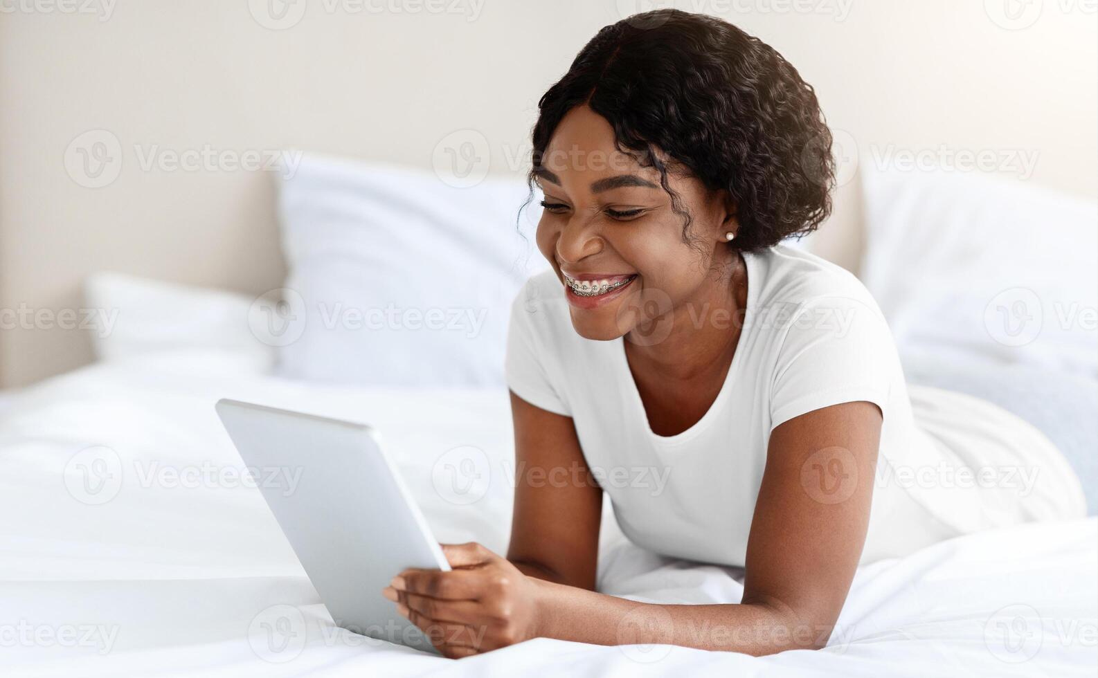 Smiling black woman laying on bed at home, using modern digital tablet, copy space. Happy african american young lady reading book or blog online, watching movie on pad, free space photo