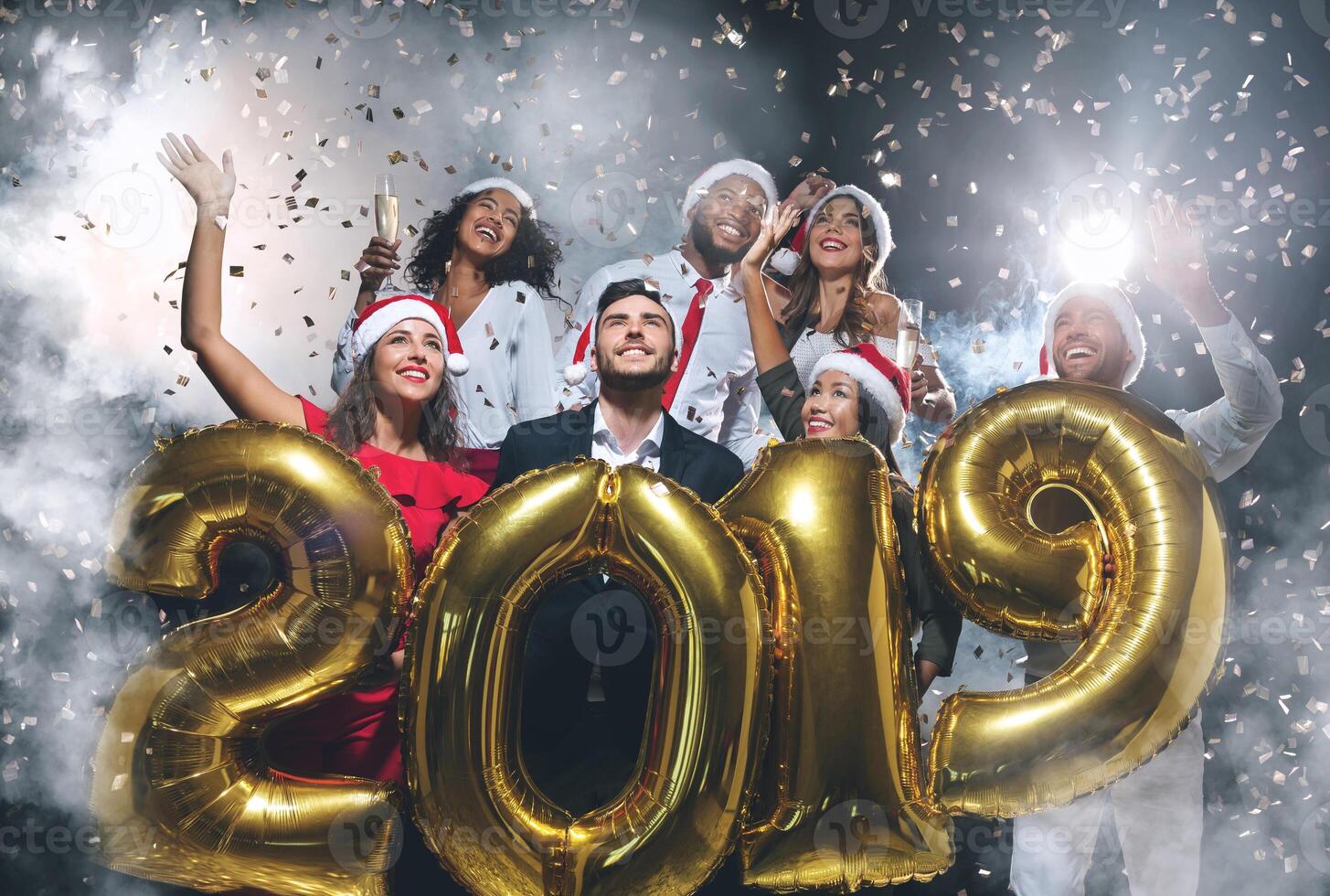Group of happy friends holding golden balloons in form of 2019 at New Year's Eve party, confetti and smoke background, copy space photo