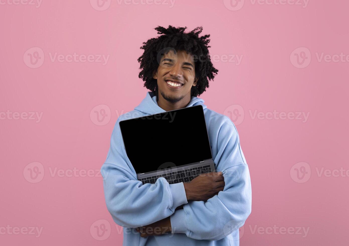 Cool African American teenager in casual wear hugging laptop pc with mockup for design on empty screen over pink studio background. Space for your website or online advertisement photo