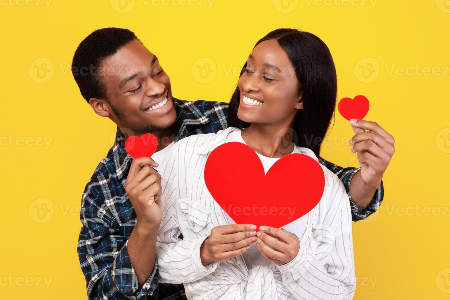 Fun together, free time and romantic celebration. Lovers generation z african american students guy and lady hold red hearts and joying, isolated on yellow background, empty space, studio shot photo