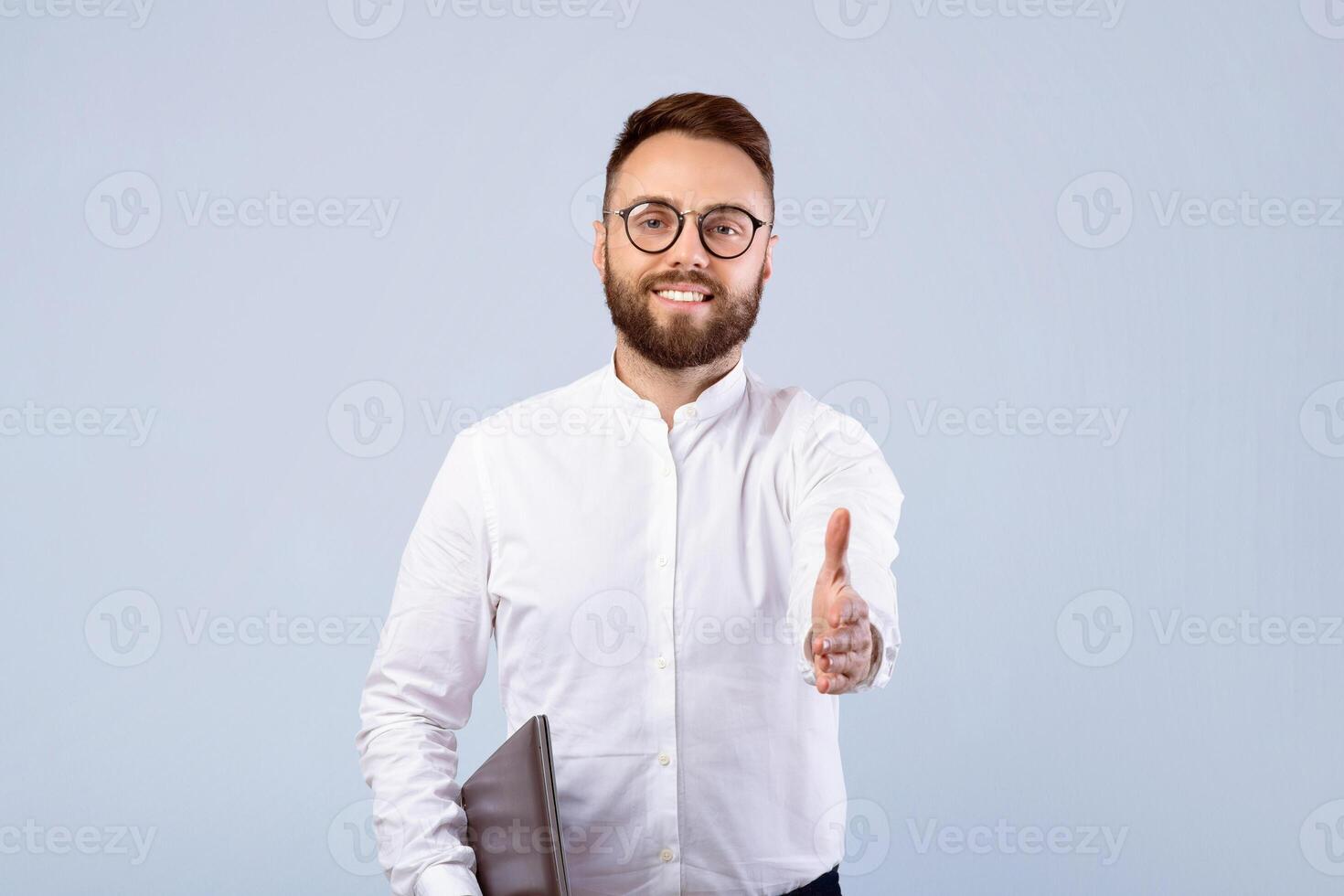 Bearded young businessman with laptop computer offering hand for handshake on grey studio background. Millennial manager with pc proposing business deal or agreement photo