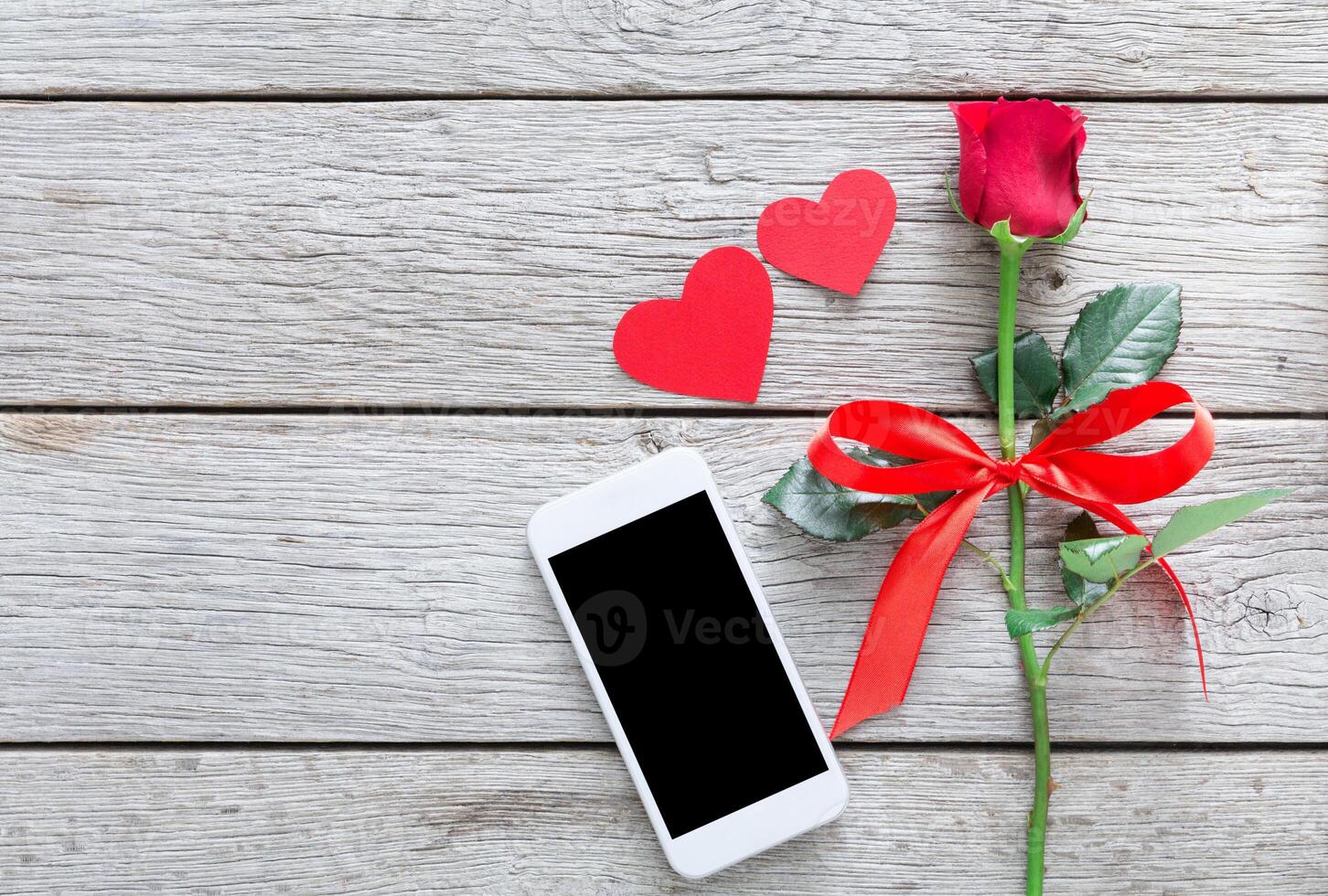 Valentine day internet sales concept, online shopping holiday background with copy space. Mobile phone on rustic wood with rose flower and hearts, top view. Card and advertising mockup photo