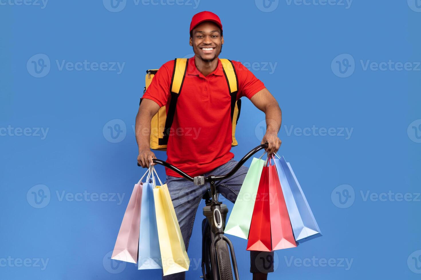 Portrait of smiling black delivery man wearing red uniform, cap and backpack riding bike and holding colorful shopping bags with customer's orders and gifts, isolated over blue studio background photo