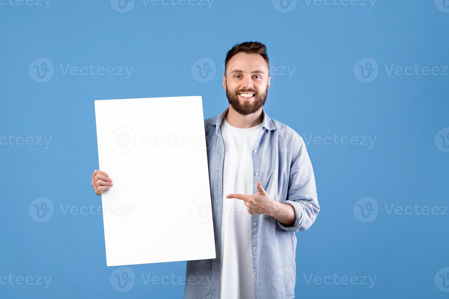 Advertising billboard. Portrait of smiling young guy pointing at blank white poster with mockup, space for your design. Happy millennial man demonstrating empty banner template photo