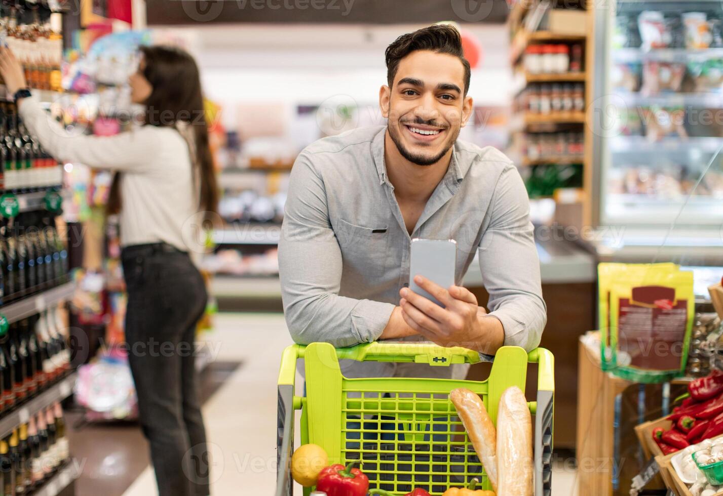 Arabic Guy With Smartphone Shopping Groceries Standing With Shop Cart Full Of Food Products In Modern Supermarket Indoor. Male Buyer Posing In Food Store On Weekend, Using Application On Cellphone photo