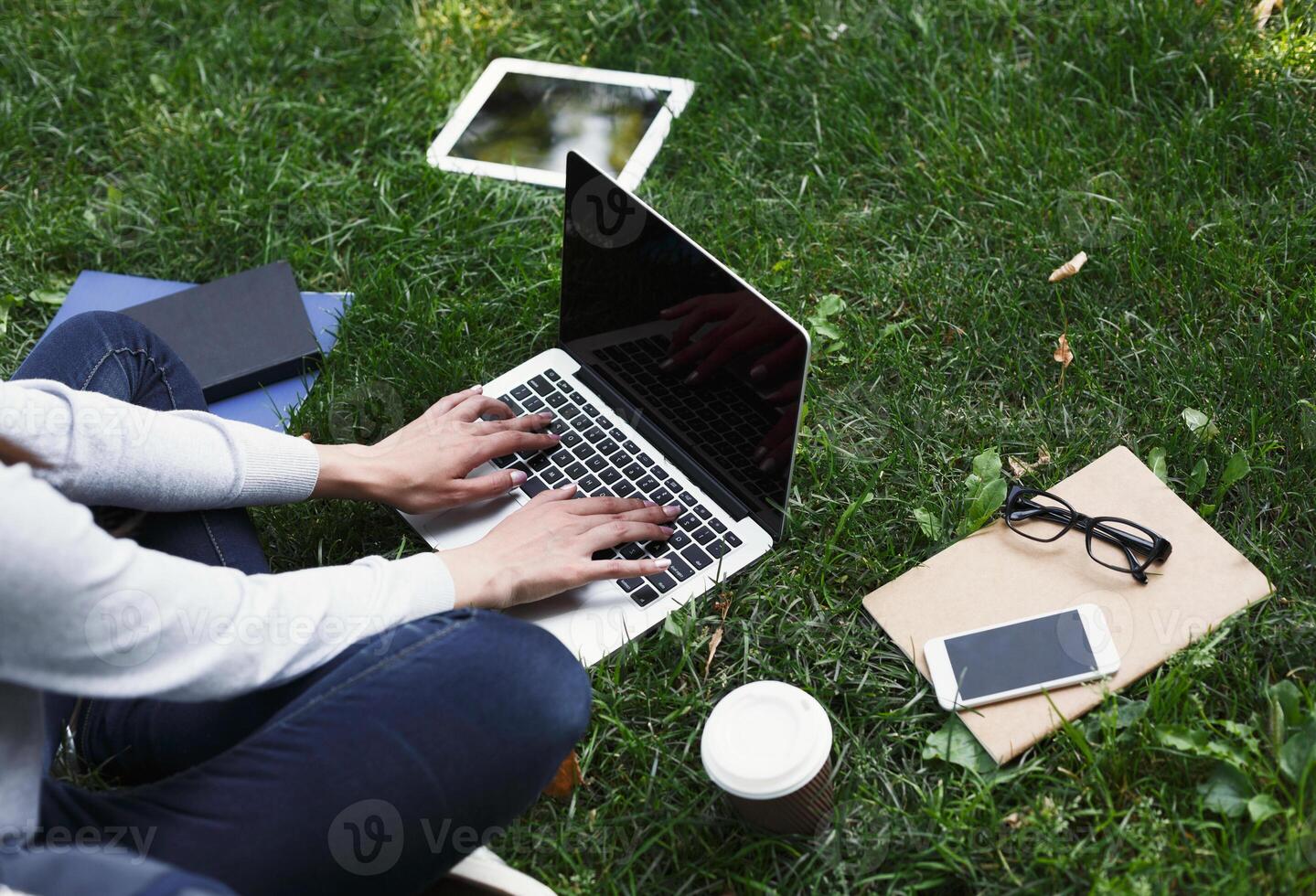 Young unrecognizable woman sitting outdoors on grass with laptop, typing, surfing internet, preparing for exams. Technology, communication, education and remote working concept, copy space photo
