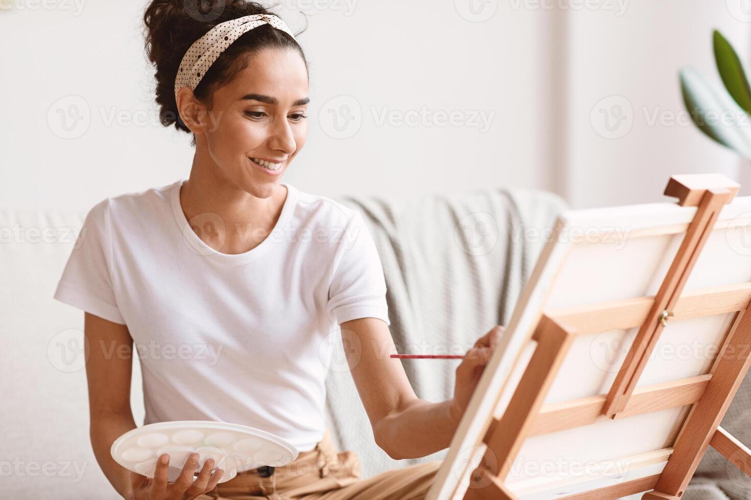 Closeup of female artist drawing at home, using easel and palette, copy space. Young brunette woman enjoying painting, spending weekend at home, having new creative hobby photo