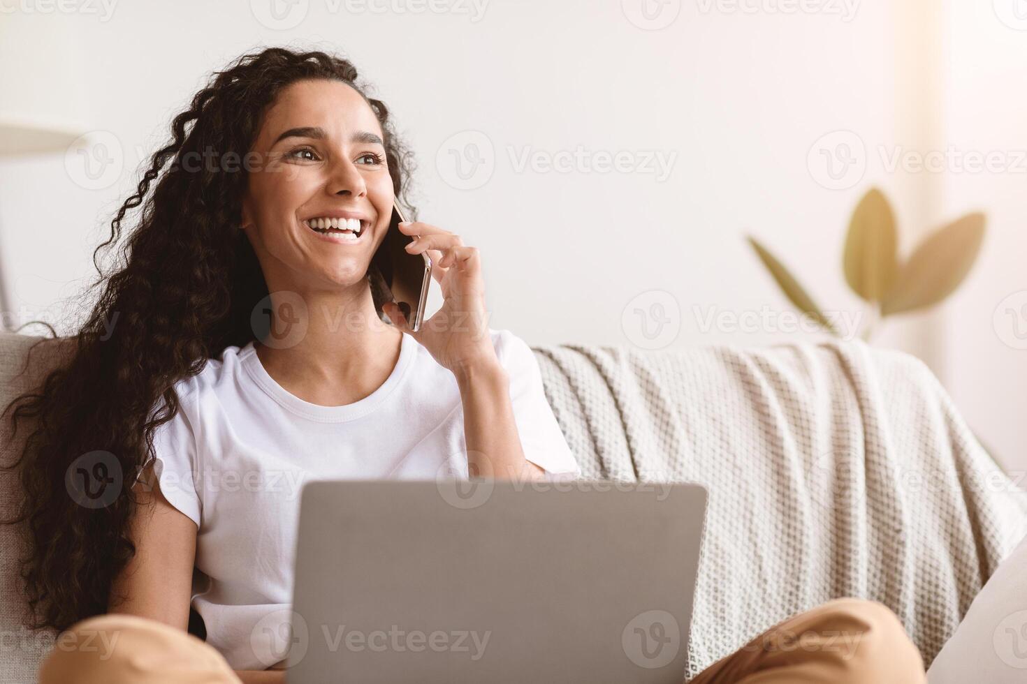 Cheerful young woman freelancer talking on mobile phone and using laptop, working from home during isolation, looking at copy space. Happy curly lady having conversation on phone photo