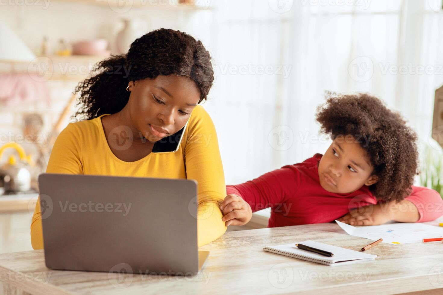 Bored Black Girl Demanding Attention From Her Busy Mom Working At Home, African American Freelancer Lady Using Laptop And Talking On Cellphone In Kitchen, Ignoring Her Lonely Child, Free Space photo