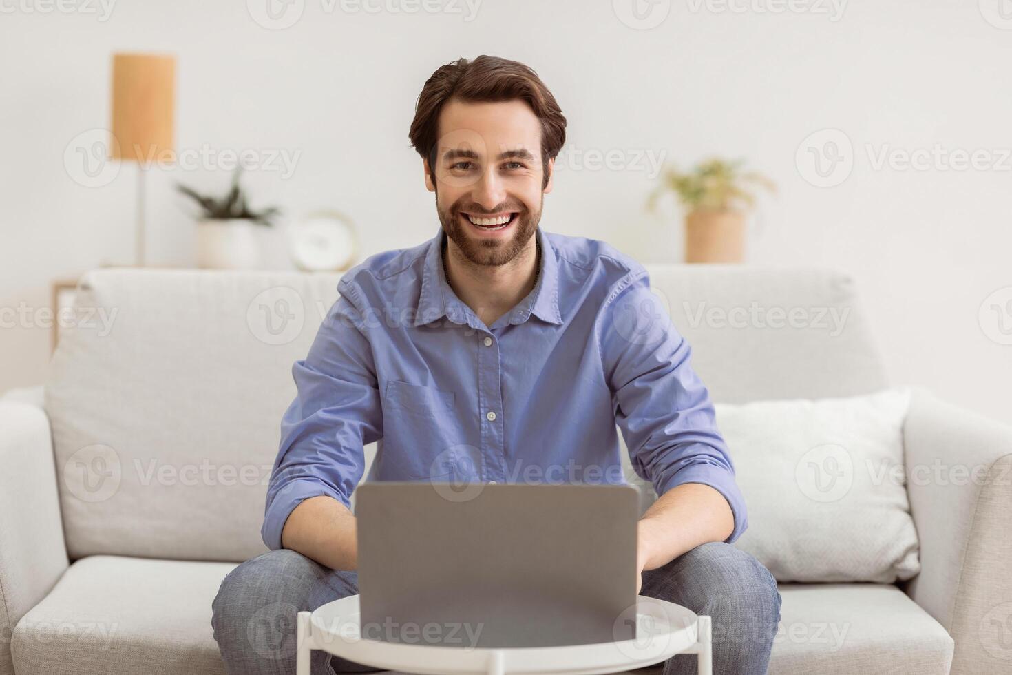 Happy man at laptop computer works online and smiles to camera sitting on couch at home. Successful remote freelancer guy working distantly indoors. Freelance career lifestyle concept photo