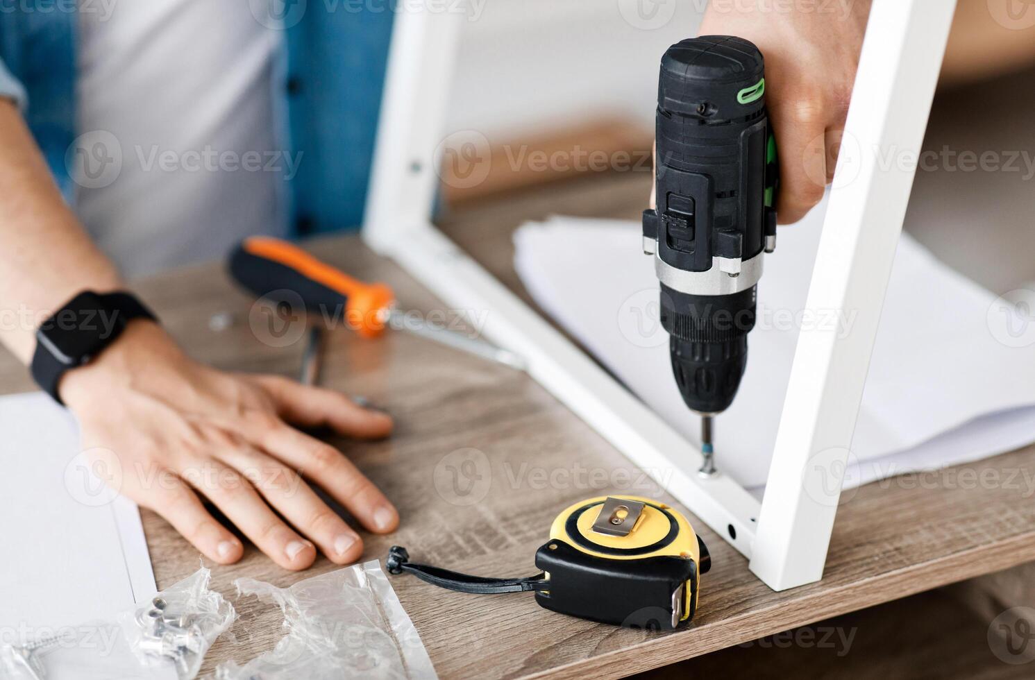 Construction worker using drill, shoots popular repair blog. Millennial man with smart watch assembles furniture with electric screwdriver and tools in interior of living room, empty space, cropped photo