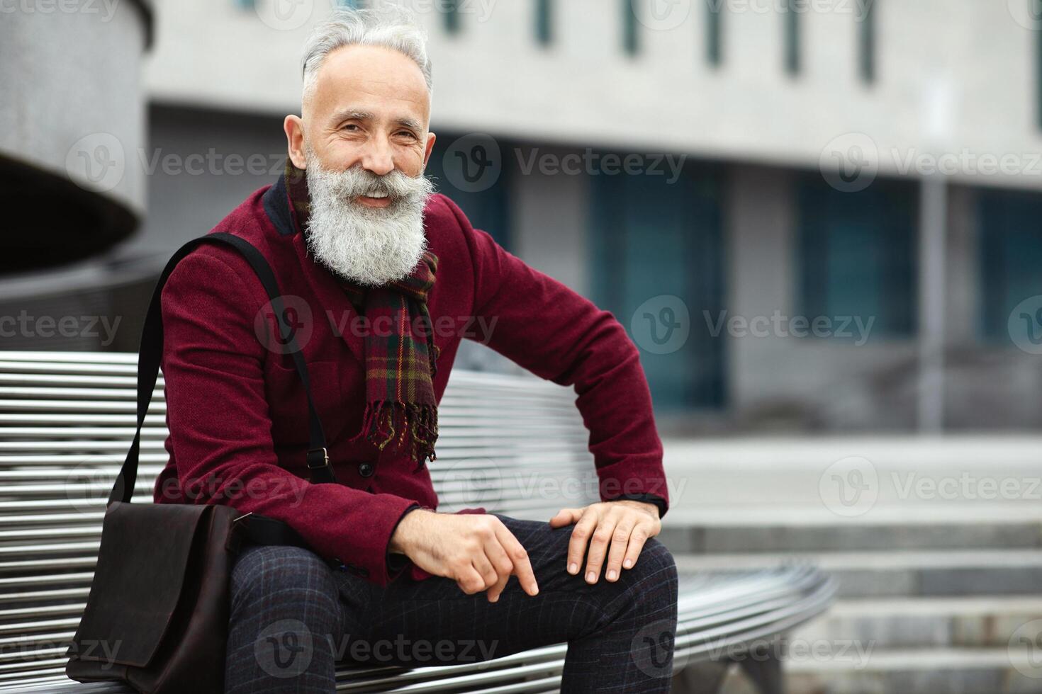 Handsome aged bearded man in stylish outfit with bag sitting on bench, smiling at camera, copy space. Happy elderly businessman enjoying his free time, breathing fresh air on the street photo