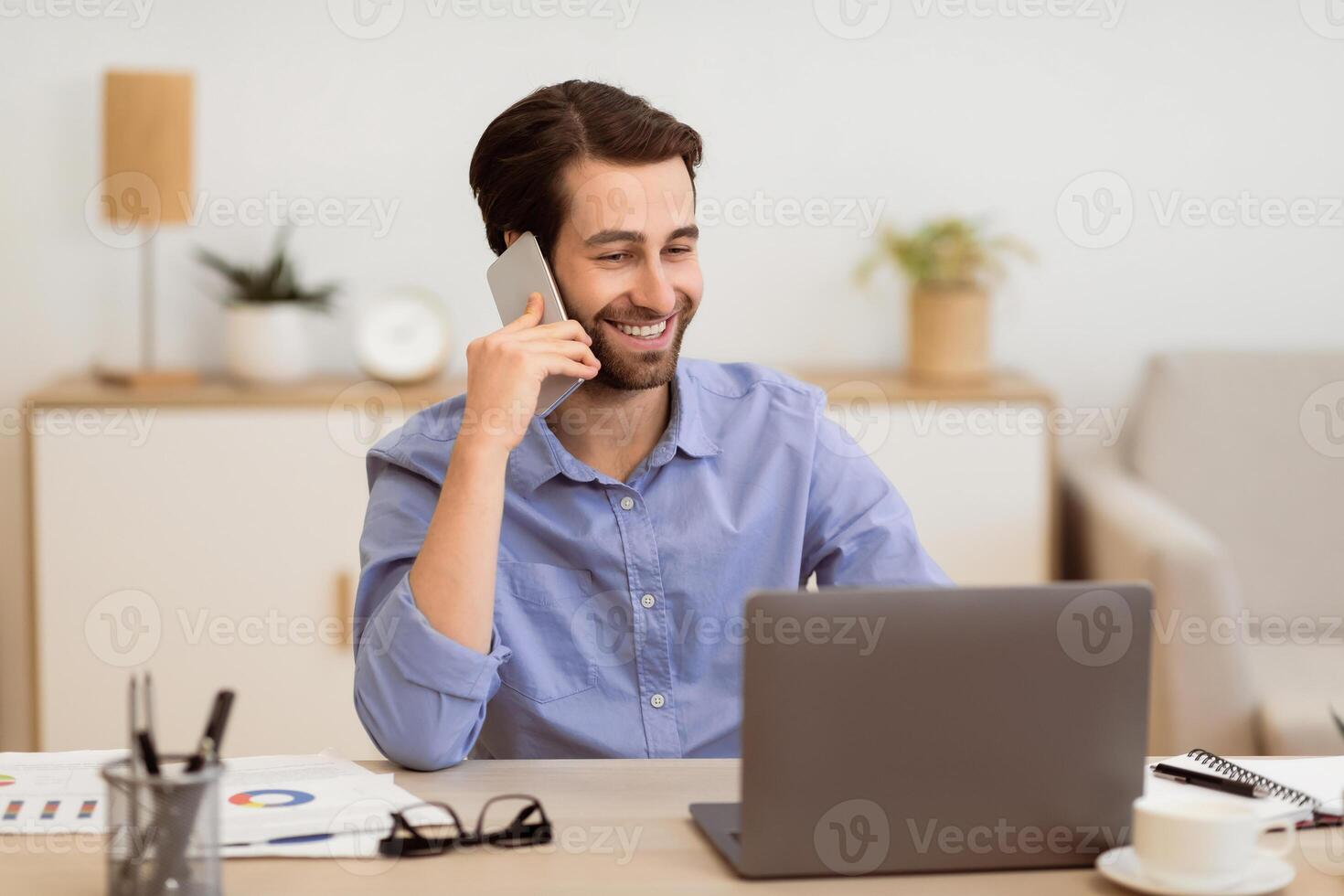 Gladful Man Talking On Phone Working On Laptop Online Sitting At Desk In Modern Office. Freelancer Guy Calling Speaking By Cellphone Indoors. Modern Mobile Communication Concept photo