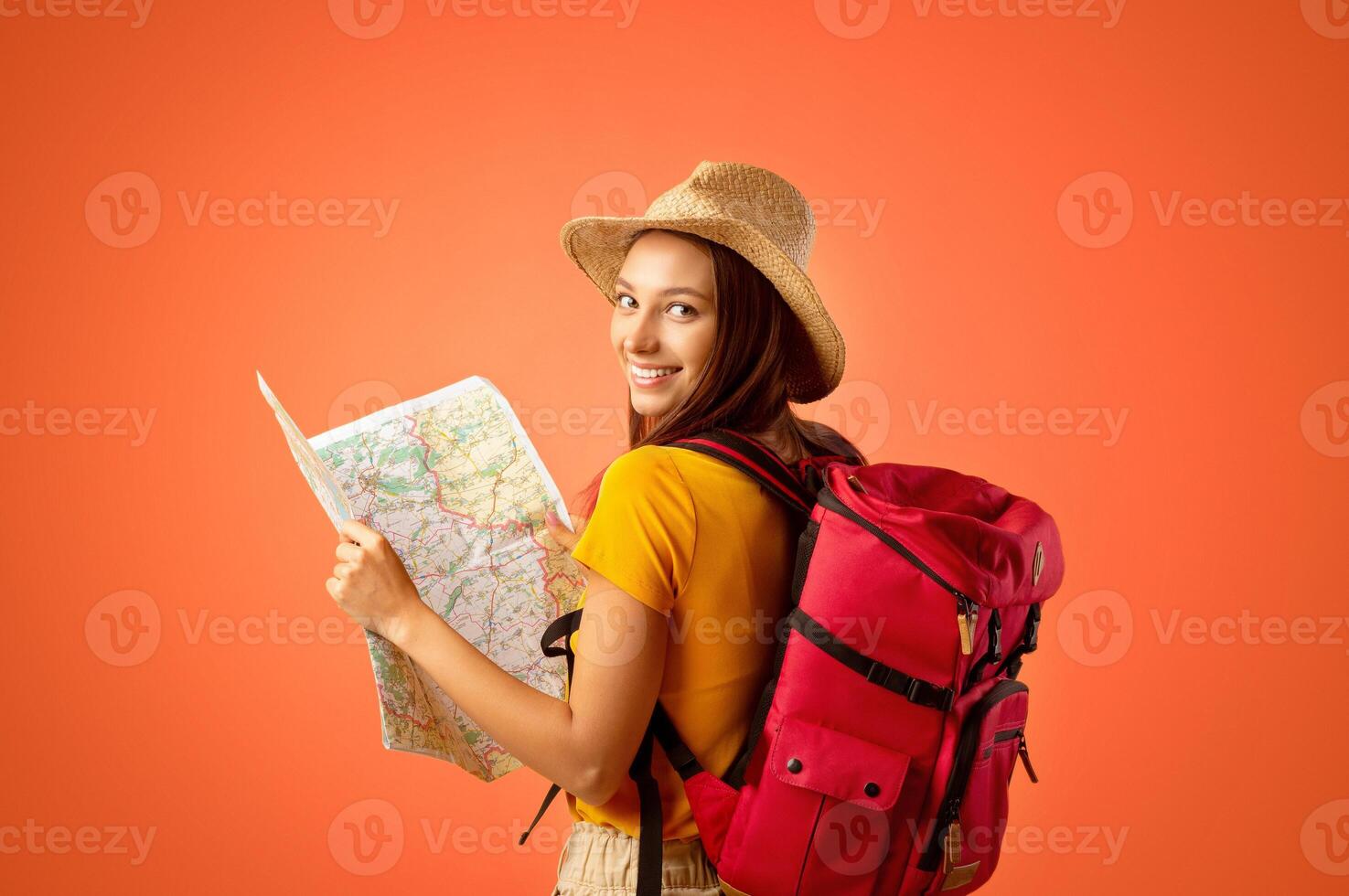 Smiling young woman tourist with backpack holding city map, yellow studio background. Cheerful lady holding map, looking over shoulder, impressed by great travelling opportunities, copy space photo