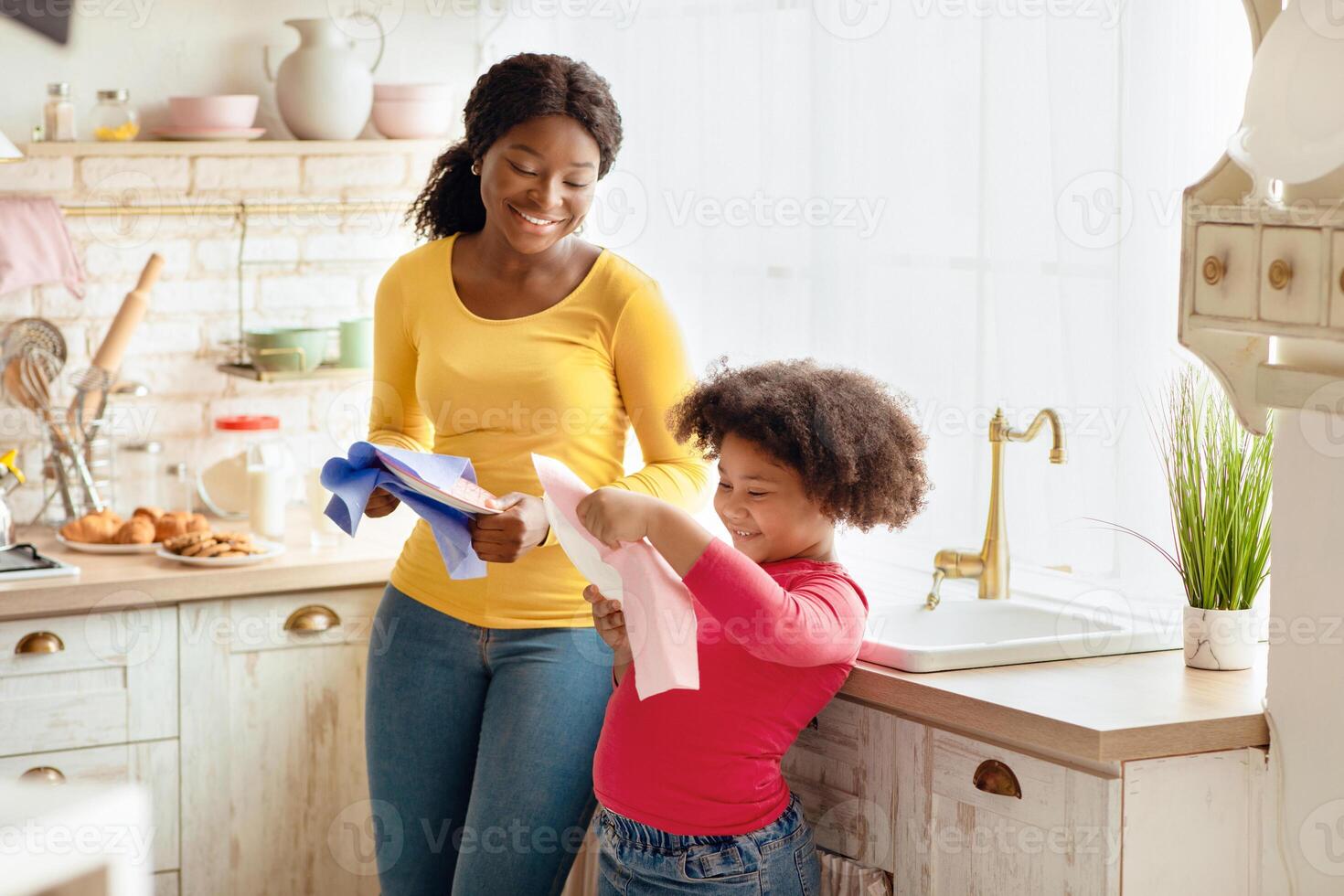 Cute African American Girl Helping Mother With Household, Wiping Dishes In Kitchen After Washing. Adorable Black Little Helper Child Enjoying Spending Time With Mom At Home, Learning Housekeeping photo