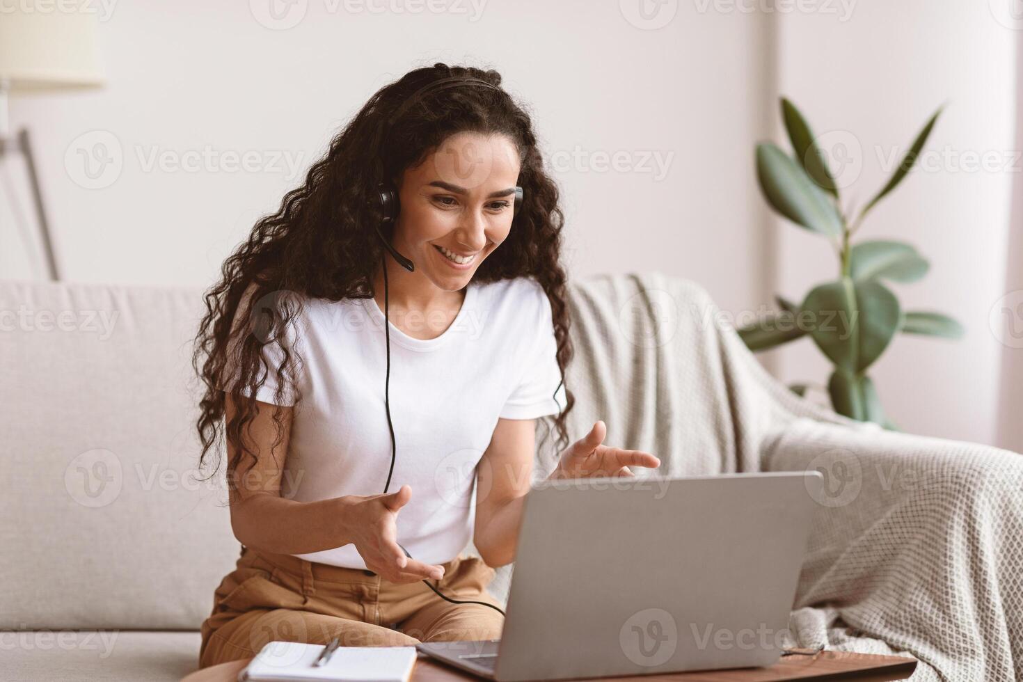 Joyful young woman with headset having online conference, working from home, copy space. Happy curly lady making call with friends or lover, using laptop, living room interior photo