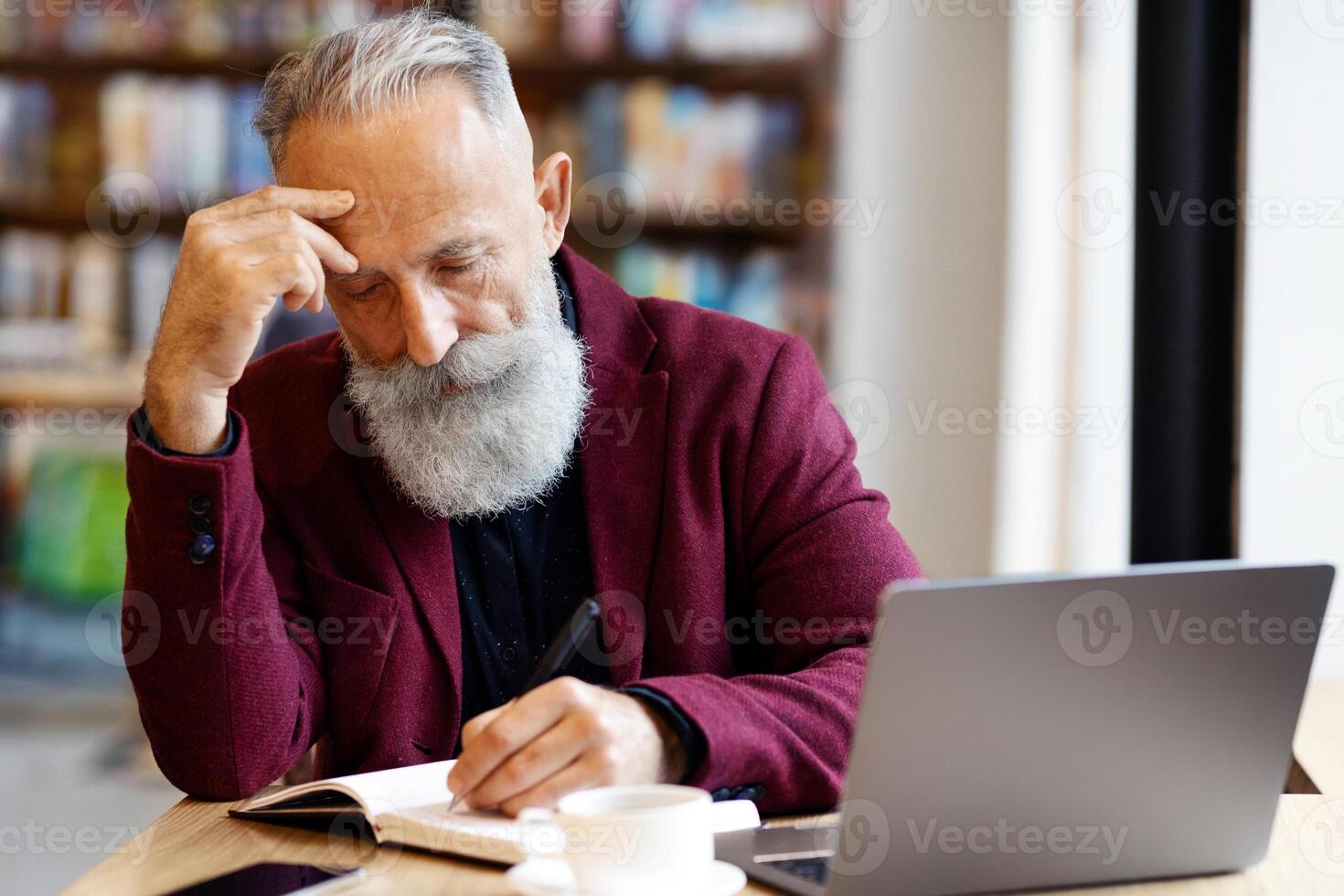 Pensive senior businessman with grey hair checking agenda, taking notes in notebook, working with laptop at cafe, copy space. Elderly concentrated man looking for job, checking his list, drinking tea photo