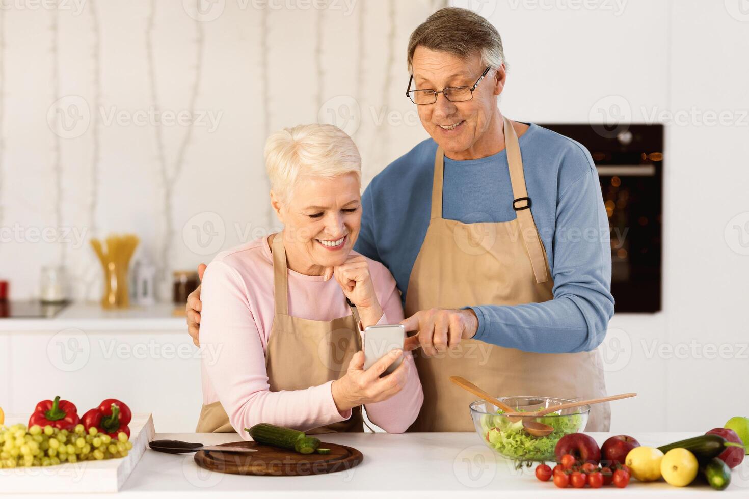 Cheerful Senior Spouses Using Cellphone Cooking Dinner Together And Browsing Internet For Online Recipes Standing In Modern Kitchen At Home. Elderly Couple Using Smartphone Preparing Meal Concept photo