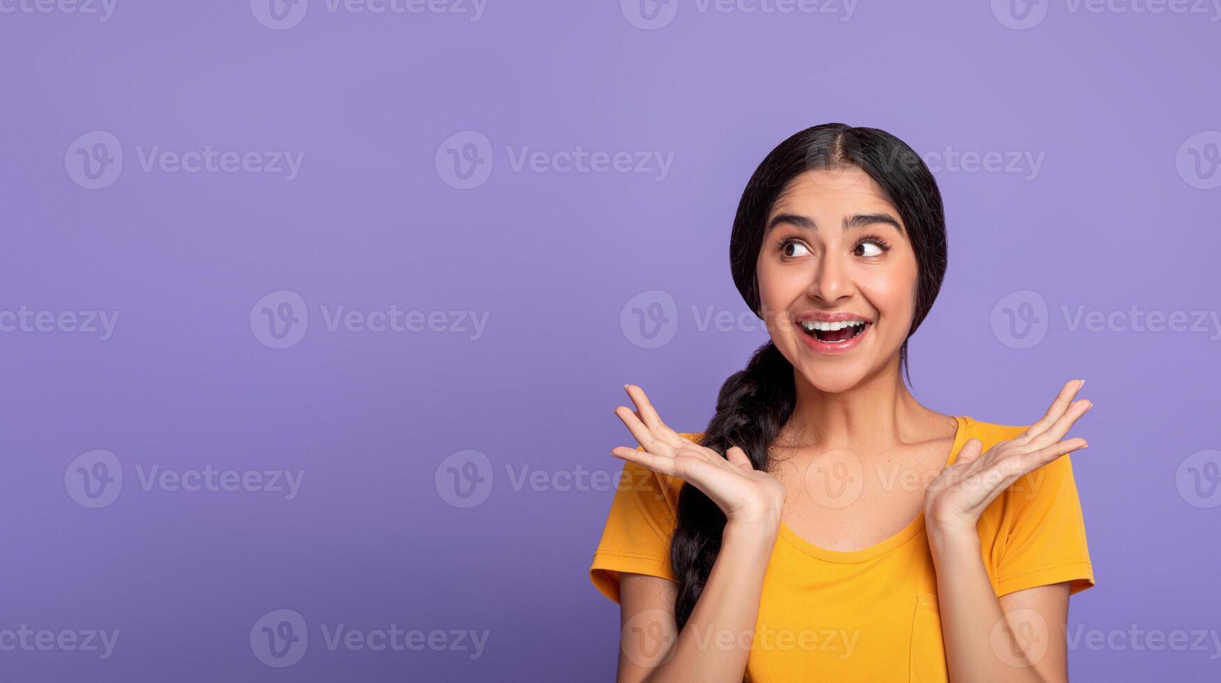 Omg. Portrait of overjoyed young indian lady spreading hands and looking aside at copy space for advertisement or promotional text, isolated over purple background, studio shot, banner photo