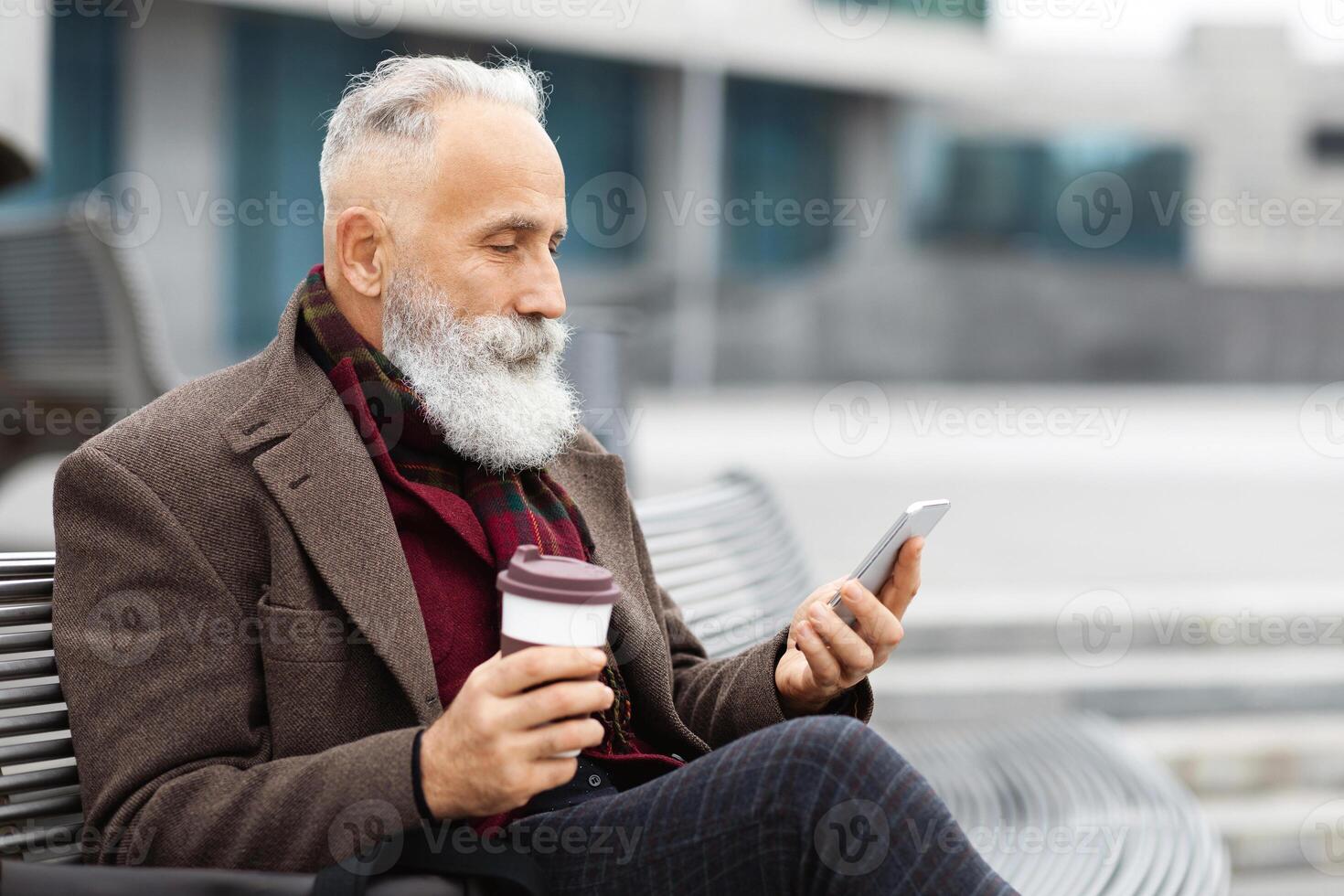 Grey-haired elderly man in warm outfit using mobile phoe and drinking coffee to go while sitting on the street, enjoying his free time outdoors, checking social media or chatting, copy space photo