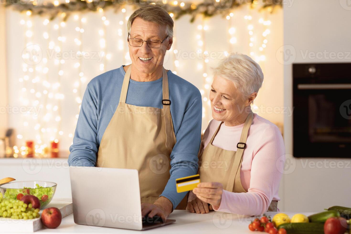 Online Grocery Shopping. Elderly Couple Using Laptop And Credit Card Buying Groceries And Ordering Food Online Standing In Modern Kitchen At Home. Happy Senior Customers Ordering Products Via Computer photo