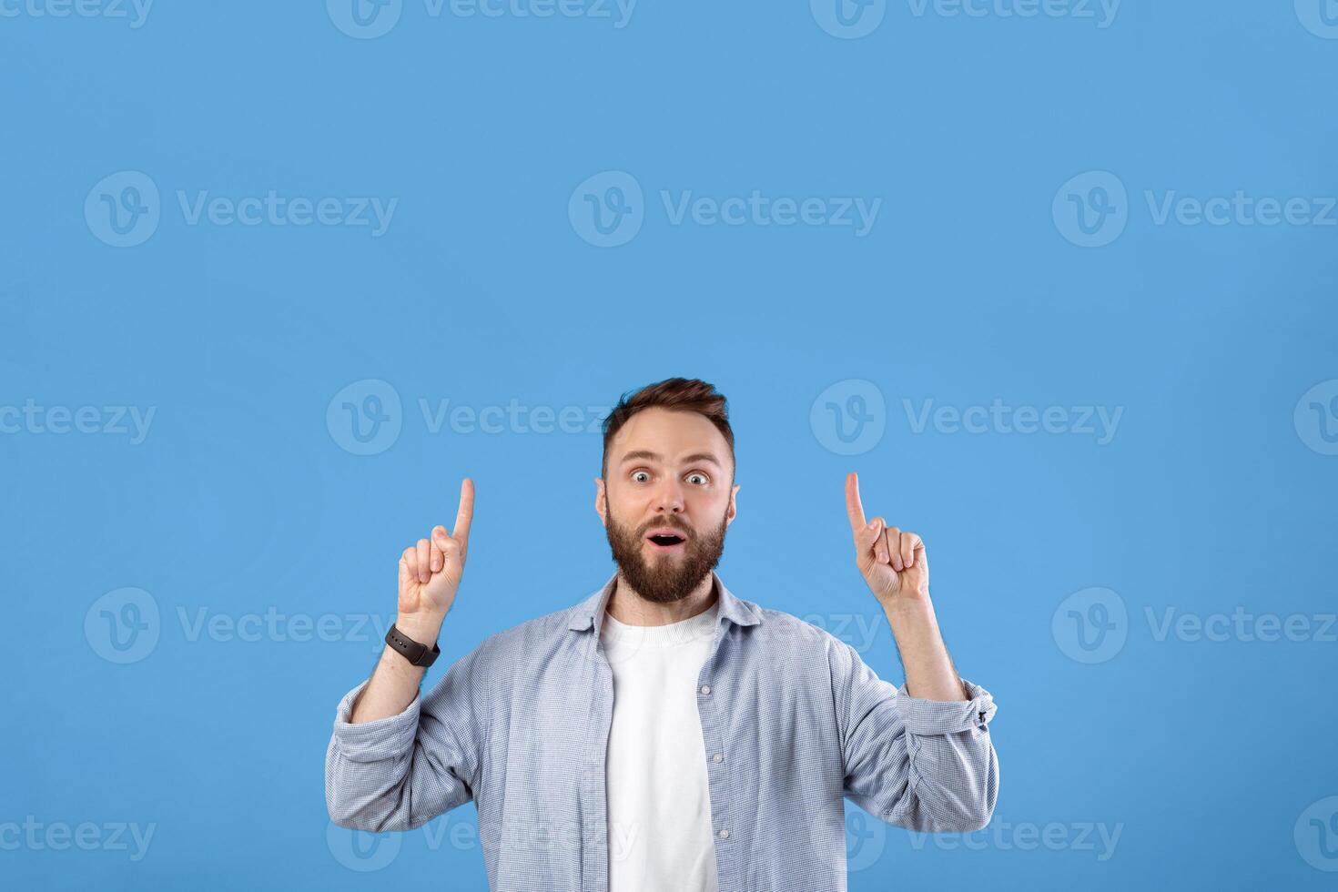Handsome bearded guy having shocked face expression, pointing at blank space over his head on blue studio background. Millennial man offering huge discounts, promoting your product photo