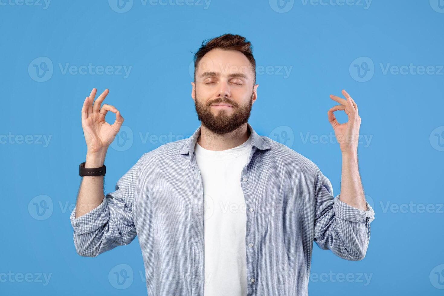 Stress management concept. Peaceful young guy meditating with closed eyes, making gyan mudra with his hands on blue studio background. Millennial man keeping calm, finding inner balance photo